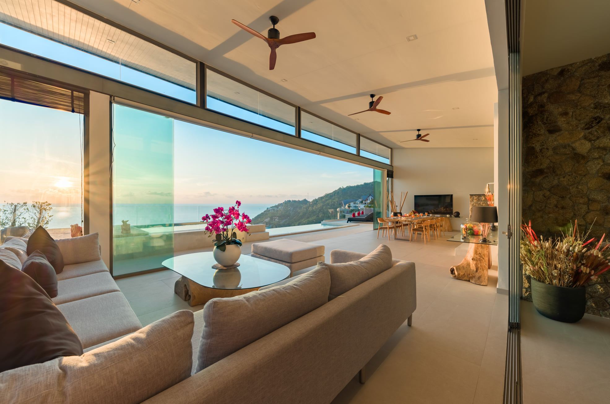 Villa 369 open-plan living room with natural stone wall and panoramic sea view at golden hour